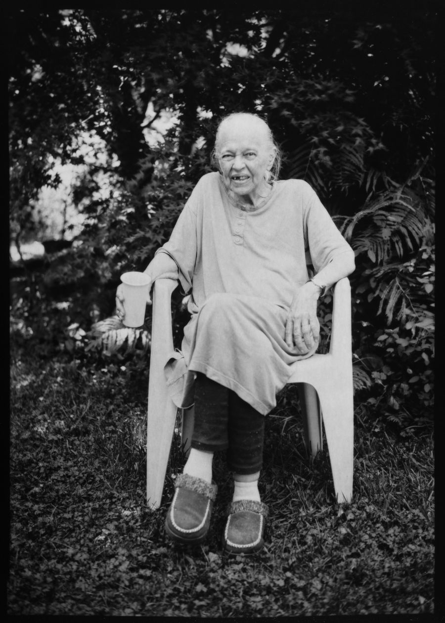 Full-length portrait of an elderly woman seated in a garden chair holding a cup, surrounded by ferns.