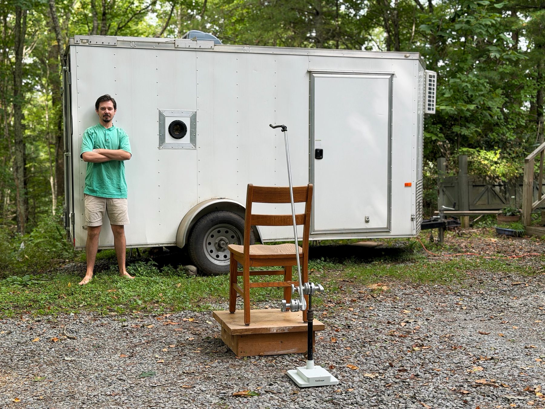 A white six-by-twelve enclosed trailer parked in the woods with a large lens mounted in its side wall. Sam Brown leans against the front of it. A wooden chair sits on a platform facing the lens.