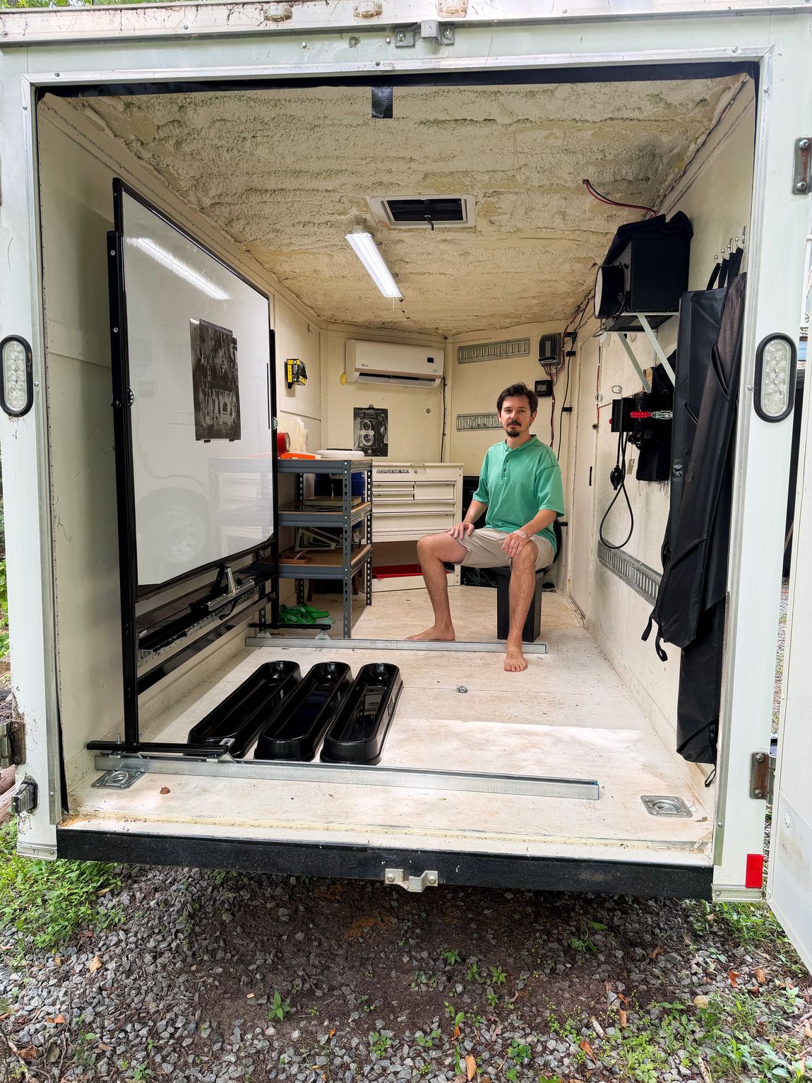The open back of the trailer showing the darkroom inside: three developing trays on the floor, a large white focusing screen, shelving, and Sam Brown seated at the far end.