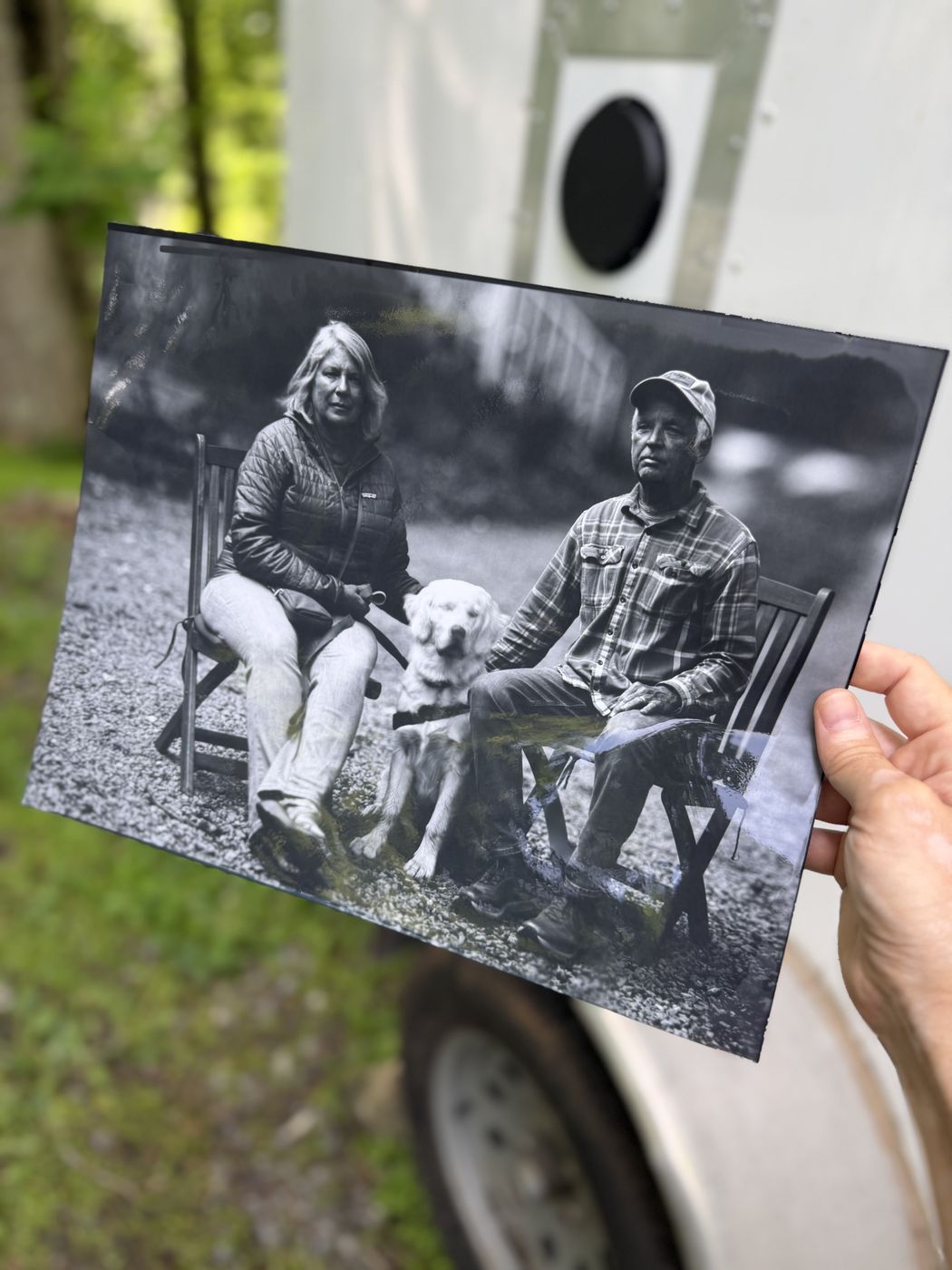 A hand holding a finished print of a couple seated with a dog, in front of the trailer with the lens visible behind.