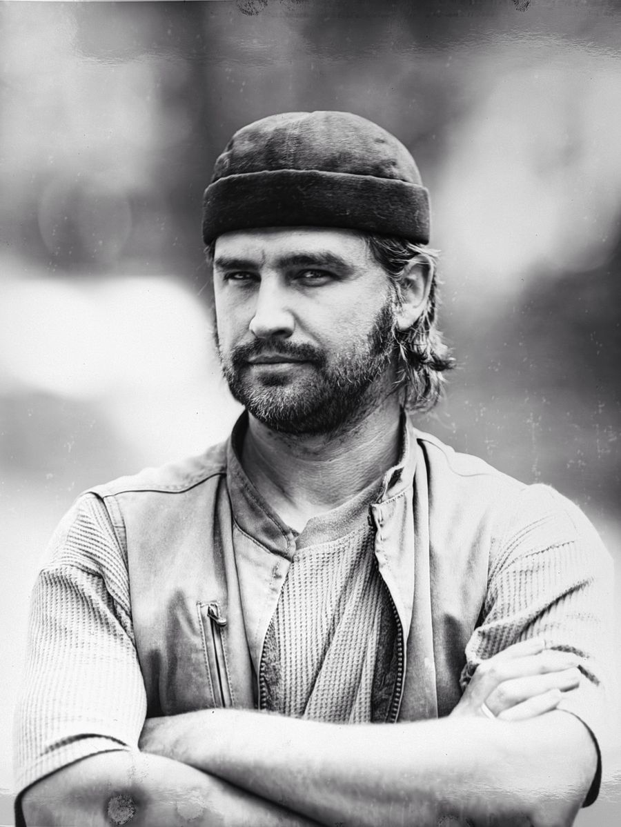 Portrait of a bearded man in a cap and work vest, arms folded.