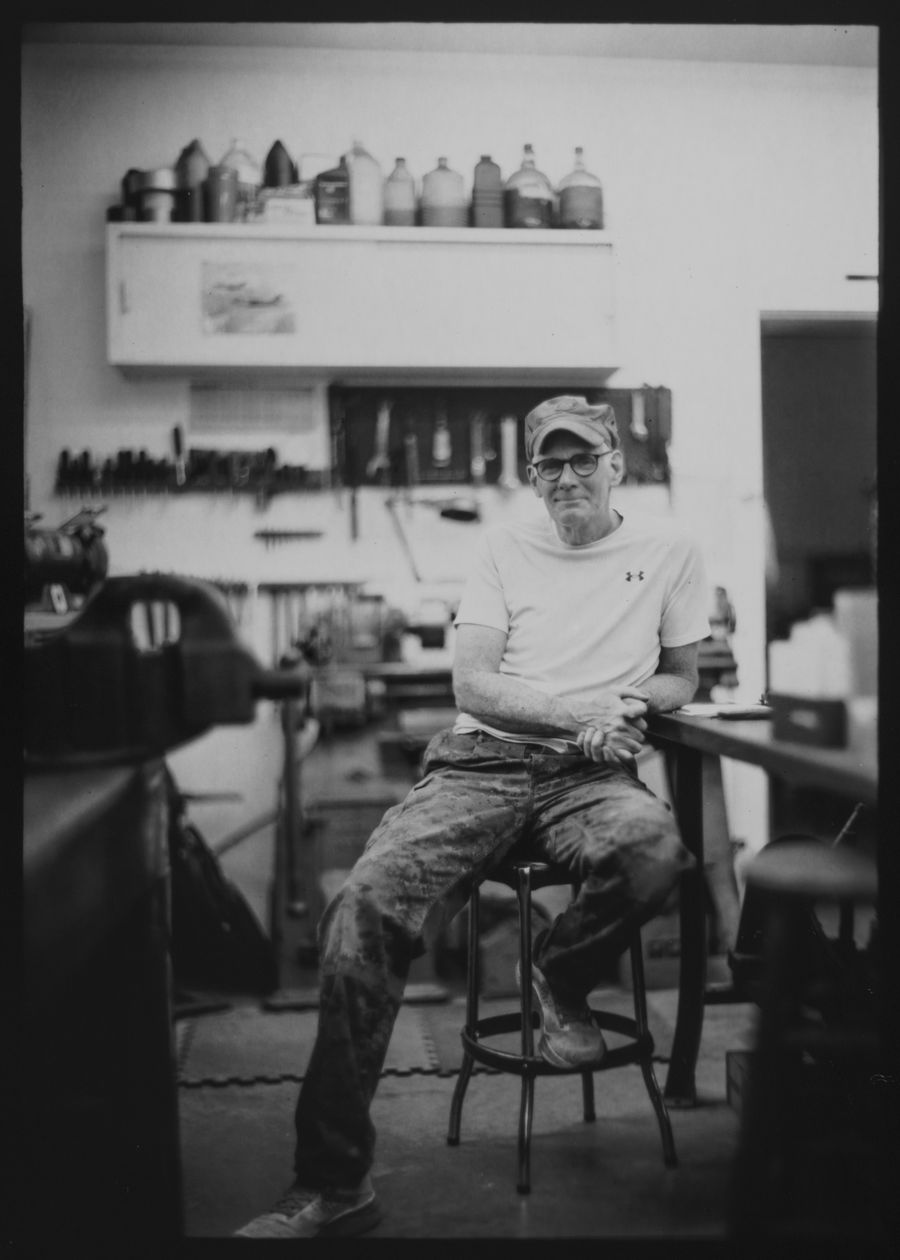Full-length portrait of a man seated on a stool in his workshop, tools racked on the wall behind him.