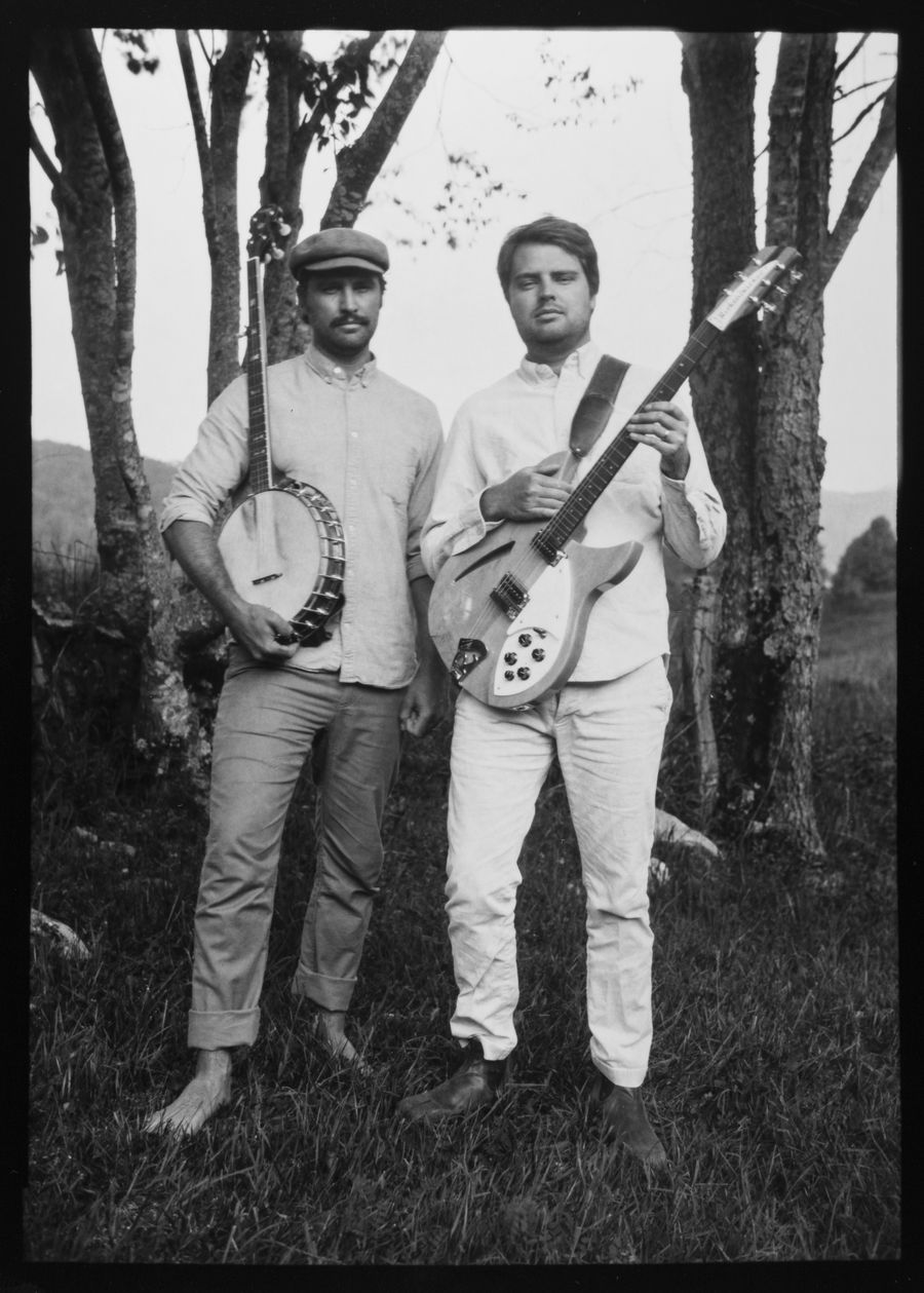 Full-length portrait of two musicians standing among trees, one holding a banjo and one an electric guitar.