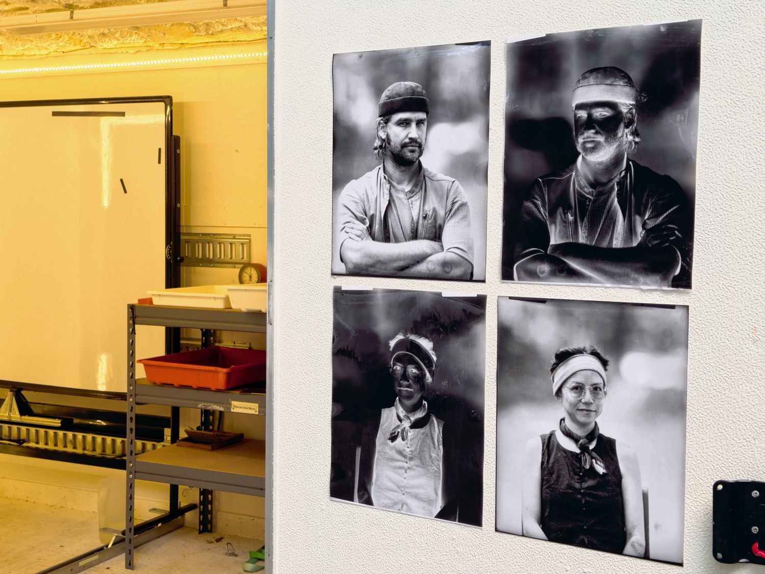 Four large prints pinned to the darkroom wall in pairs, each negative beside its finished positive, with the safelit darkroom visible alongside.